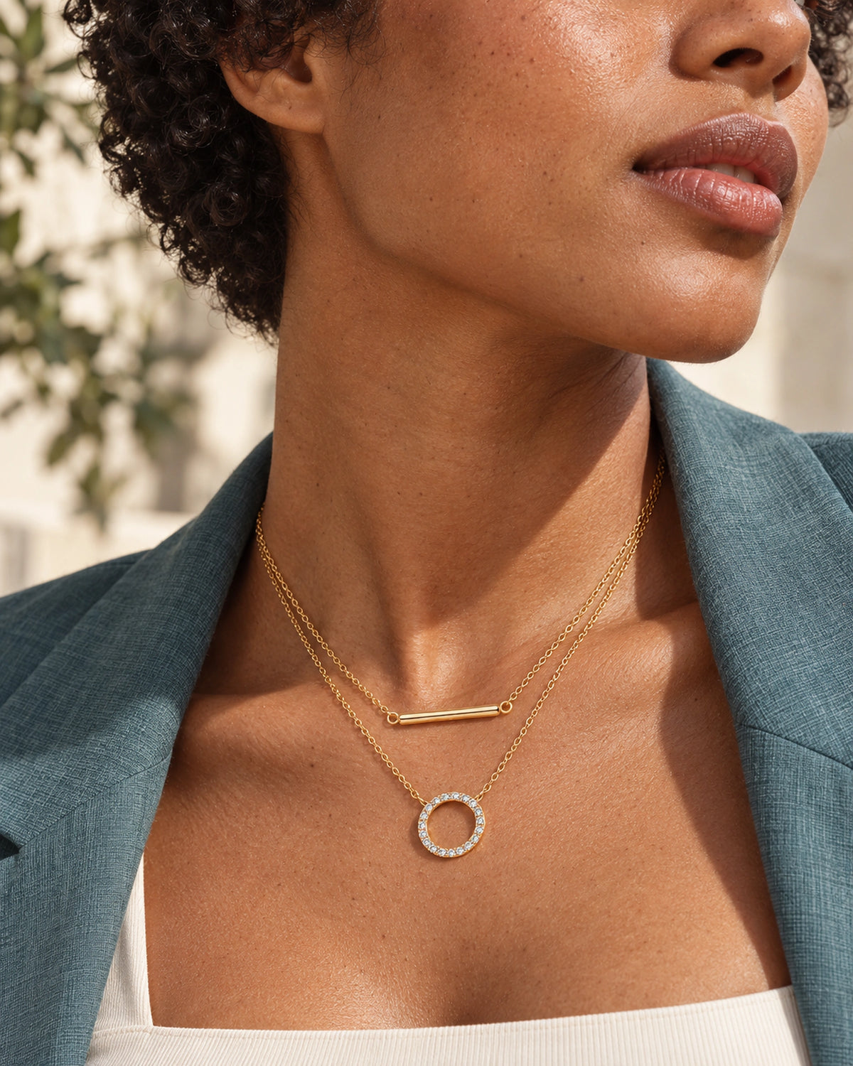 Simone gold bar and open-circle moissanite layered necklace worn with a teal blazer and ivory top in natural sunlight.